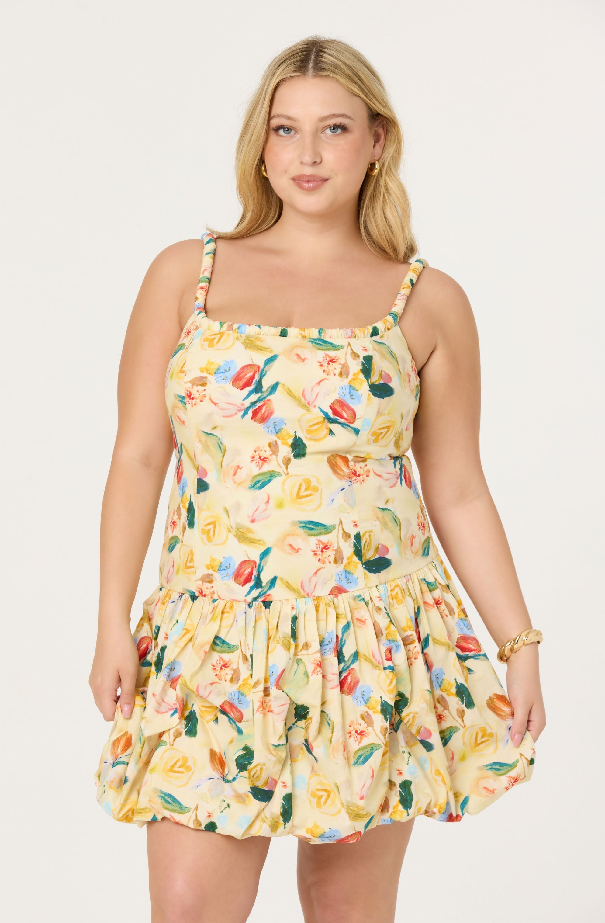 A woman is wearing a sleeveless, knee-length dress with spaghetti straps. The dress features a colorful, abstract floral pattern on a light, creamy yellow background. The skirt part of the dress is gathered and ruffled, giving it a voluminous and playful appearance.
