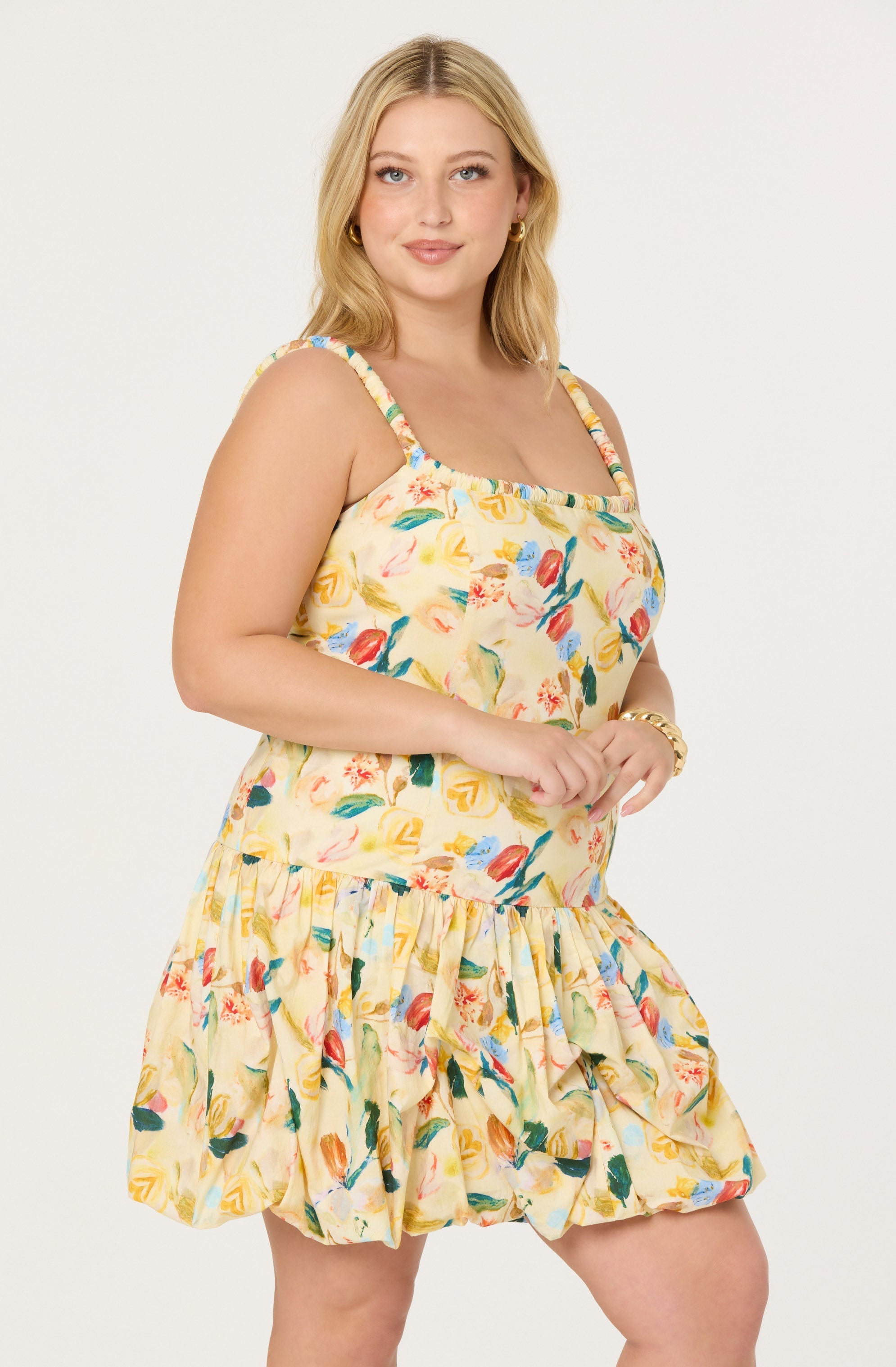 A woman is wearing a sleeveless dress with a colorful floral pattern on a light yellow background. The dress has thin, multicolored strap details on the shoulders and a ruffled hem.