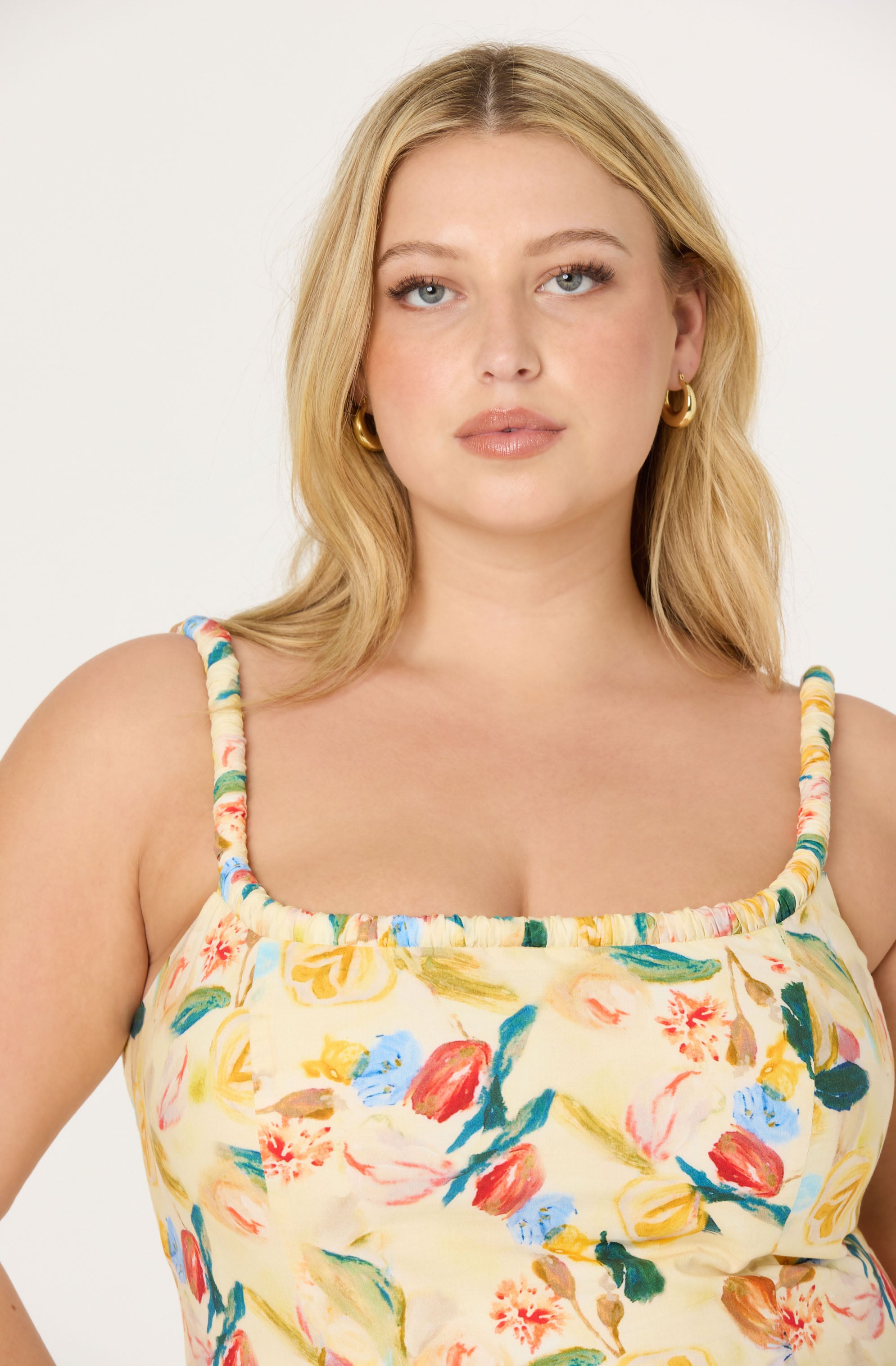 A woman is wearing a sleeveless, floral-print top with colorful flowers in shades of red, pink, yellow, blue, and green on a light, creamy background. The top has braided straps and a gathered neckline. She accessorizes with gold hoop earrings.