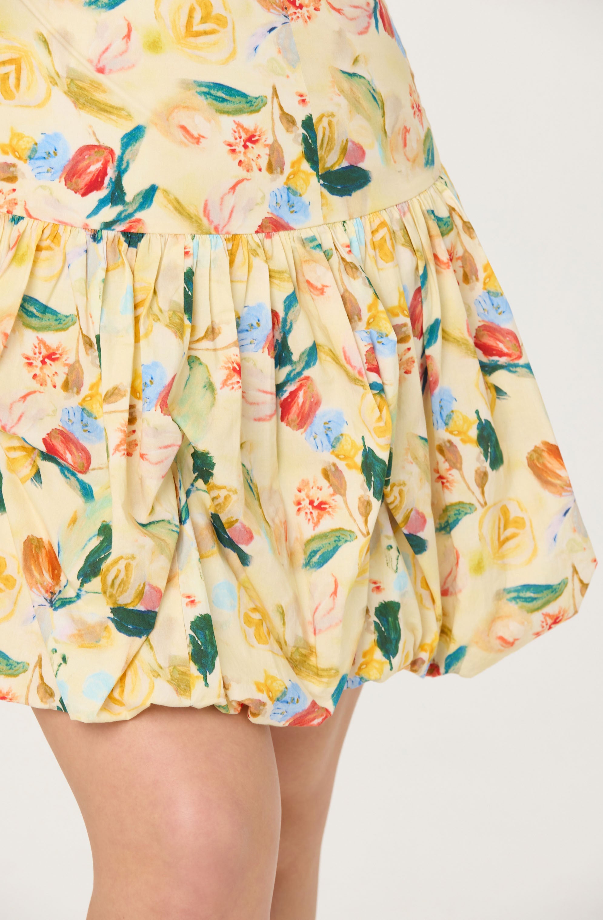 A close-up of a dress with a floral pattern featuring various colors, including yellow, red, blue, green, and pink. The fabric is light and appears to be soft, with gathering at the waist creating a voluminous, bubble-style skirt. The dress has a smooth, rounded waistband and the skirt is slightly puffed at the bottom.