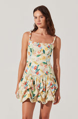 A young woman is wearing a sleeveless, knee-length dress with wide, ruffled straps. The dress features a colorful, abstract floral print on a light background, with a fitted bodice and a gathered, voluminous skirt that has a bubble hem.