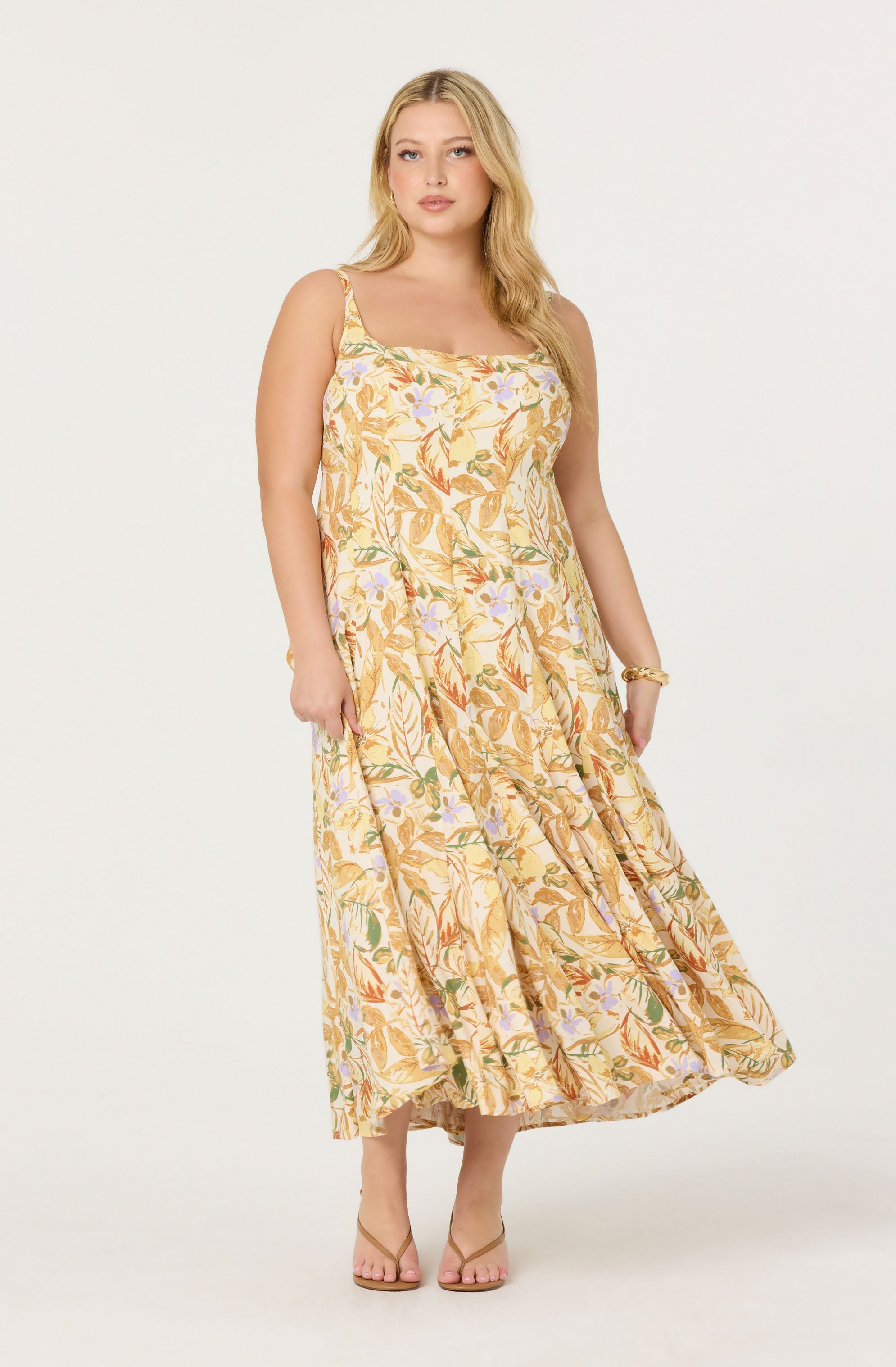 A woman is wearing a sleeveless, ankle-length sundress with thin straps. The dress features a light, floral pattern with shades of yellow, orange, green, and hints of purple, creating a soft, summery look. She is also wearing a gold bracelet on her right wrist and beige open-toe sandals.