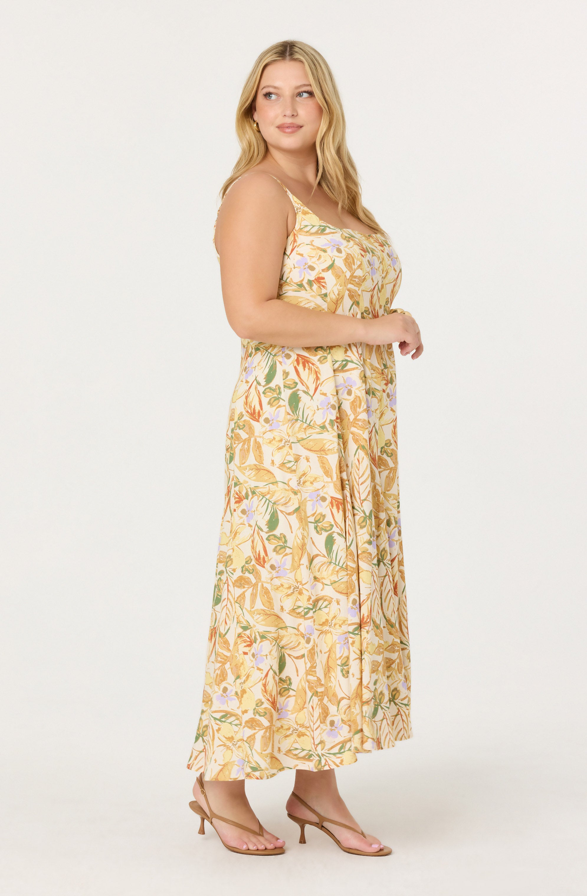 A woman is wearing a long, sleeveless, flowy dress with a pastel-colored floral and leaf pattern. She is also wearing open-toe heels in a nude shade.