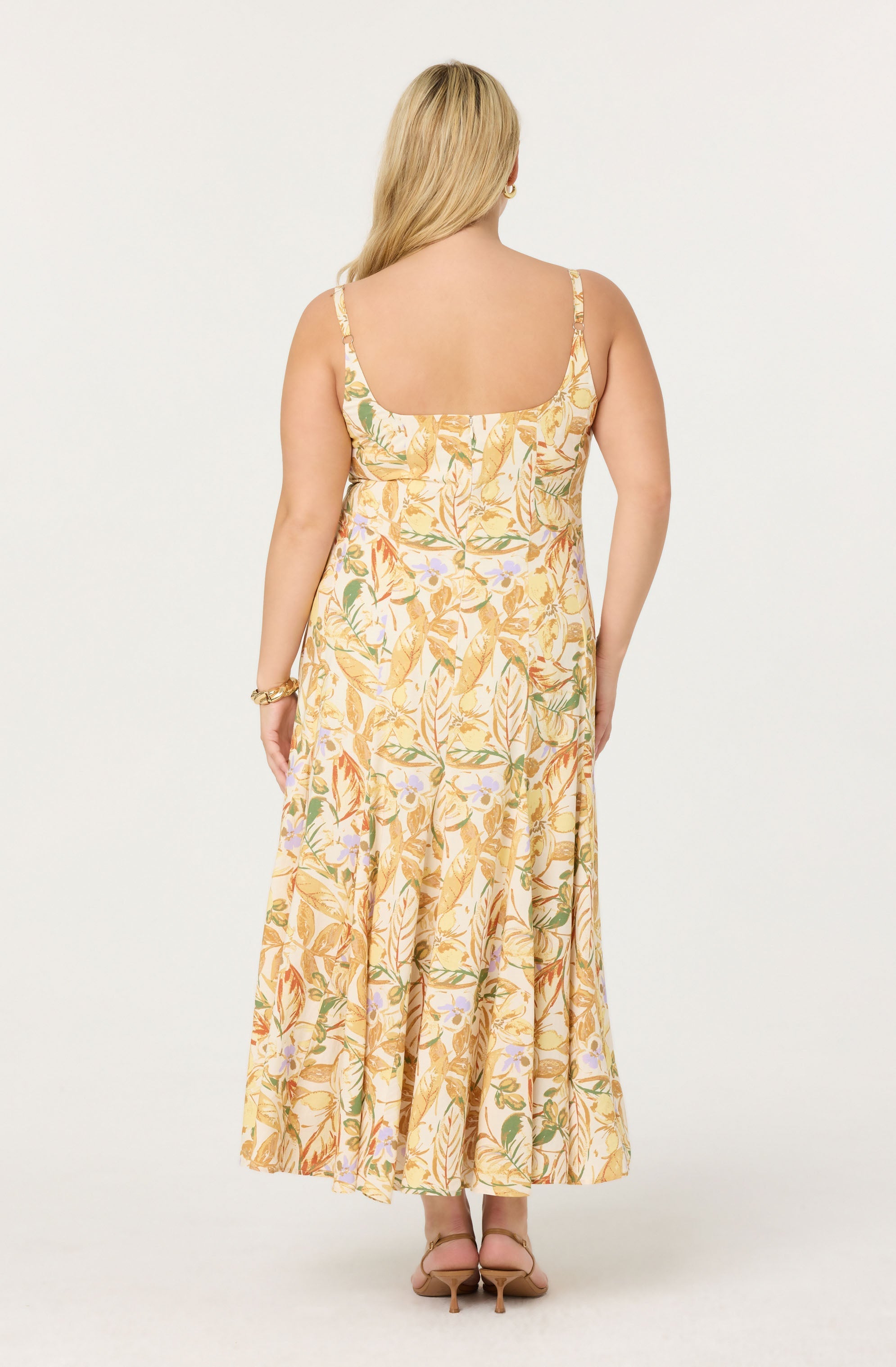 A woman is wearing a long, sleeveless, flowy dress with a floral pattern. The dress features wide shoulder straps and a square neckline. The fabric has a light, pastel color palette with shades of yellow, green, orange, and purple in a botanical print. She is also wearing tan high-heeled sandals and a gold bracelet on her left wrist.