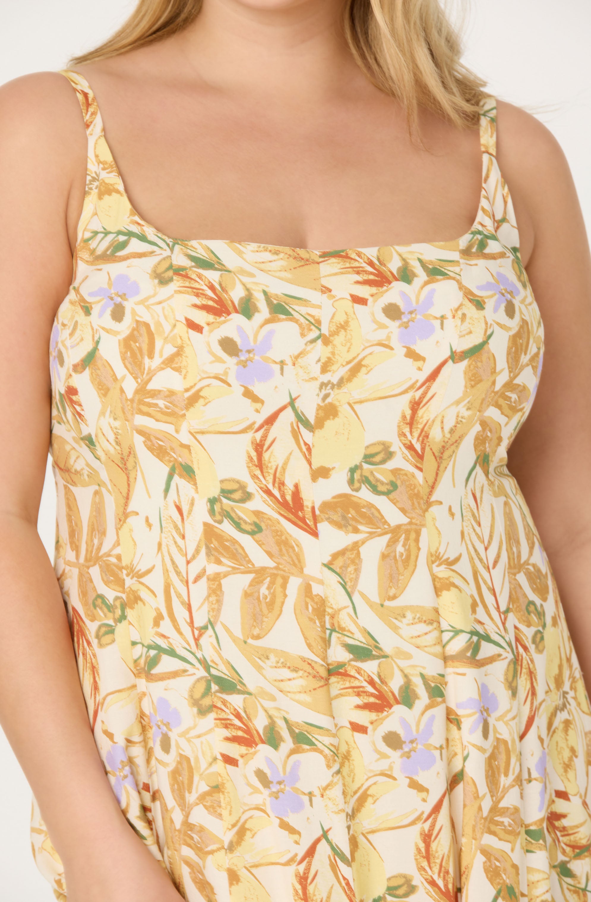A woman is wearing a sleeveless dress with spaghetti straps featuring a tropical floral pattern. The dress has a square neckline and is decorated with yellow, green, orange, and light purple floral and leaf designs on a light background.