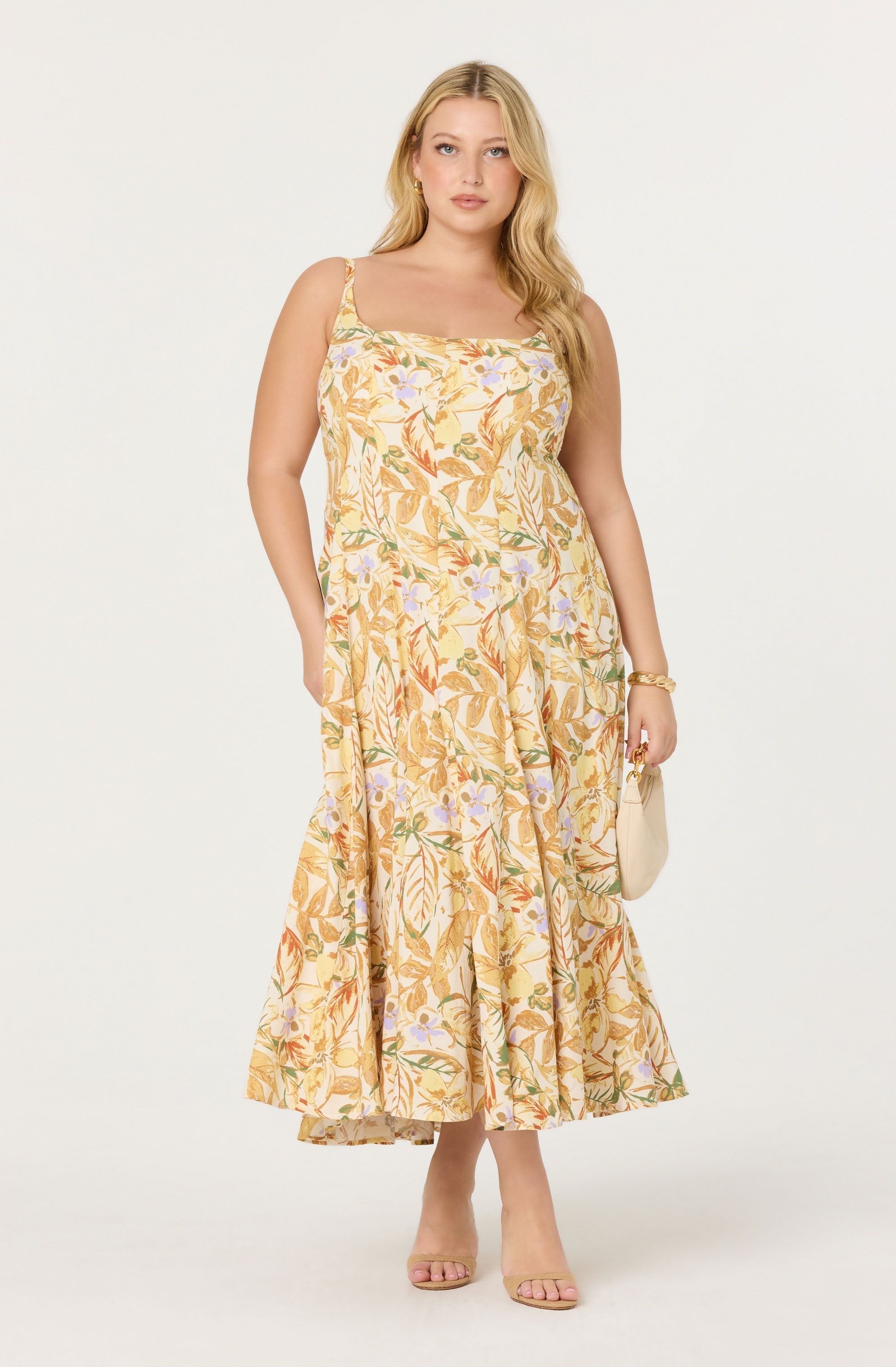 A woman is wearing a sleeveless, ankle-length dress with a floral print in shades of yellow, orange, green, and purple. The dress has thin straps and a fitted bodice with a gathered, flowing skirt. She accessorizes with a gold bracelet, a small beige handbag, and nude open-toe heels.