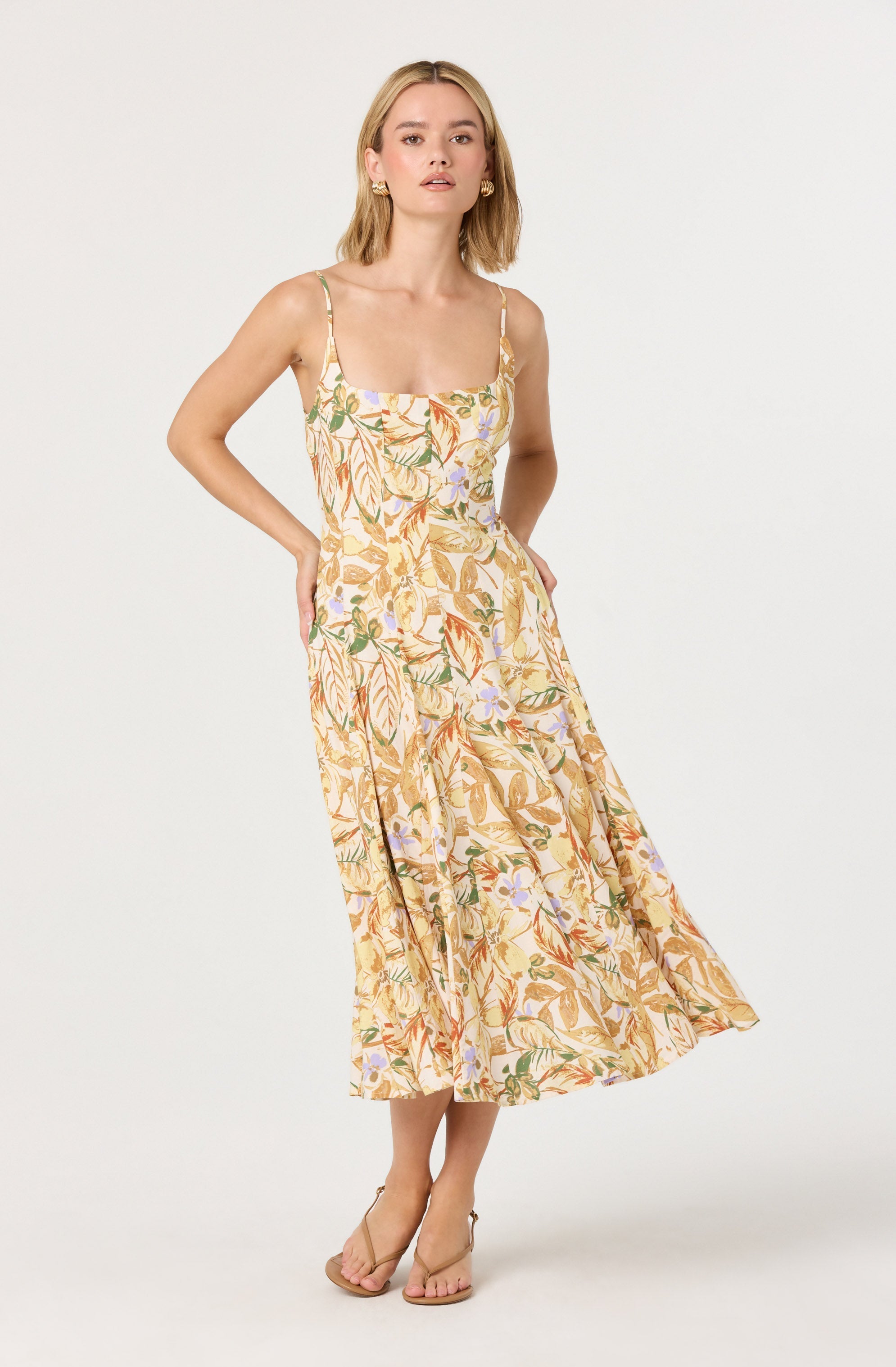 A woman is wearing a sleeveless, ankle-length dress with a fitted bodice and thin spaghetti straps. The dress features a light background with a colorful floral and leaf pattern in shades of orange, green, purple, and beige. She is also wearing beige open-toe high-heeled sandals.