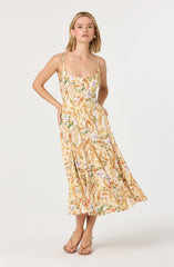 A woman is wearing a sleeveless, ankle-length dress with a fitted bodice and thin spaghetti straps. The dress features a light background with a colorful floral and leaf pattern in shades of orange, green, purple, and beige. She is also wearing beige open-toe high-heeled sandals.