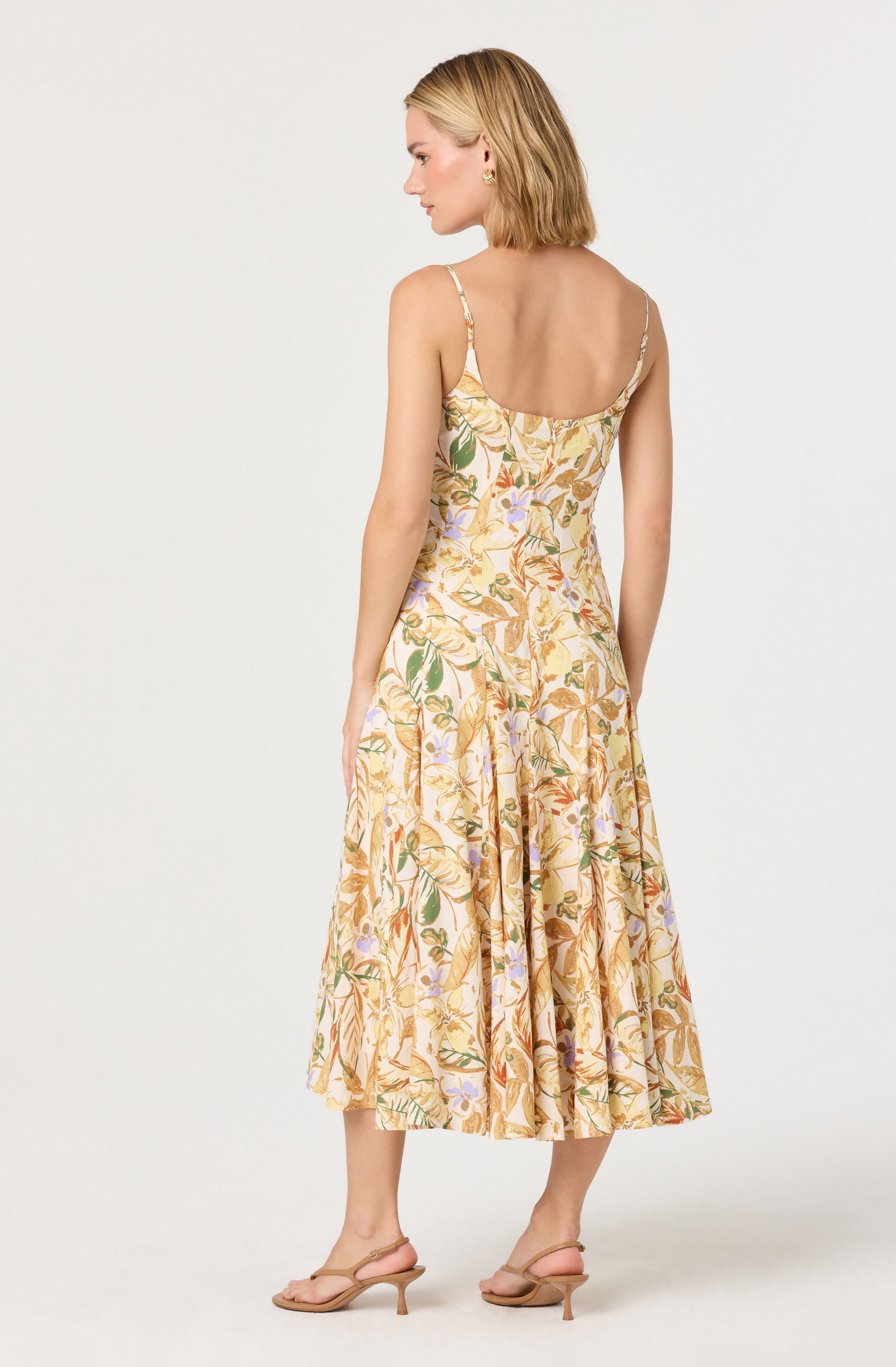 A woman is wearing a sleeveless, spaghetti strap dress with a floral pattern in shades of yellow, orange, green, and light purple. The dress has a fitted bodice and a flowing, mid-length skirt. She is also wearing beige, open-toe, low-heeled sandals.