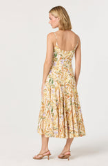A woman is wearing a sleeveless, spaghetti strap dress with a floral pattern in shades of yellow, orange, green, and light purple. The dress has a fitted bodice and a flowing, mid-length skirt. She is also wearing beige, open-toe, low-heeled sandals.