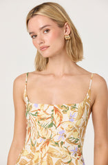A woman is wearing a sleeveless dress with thin spaghetti straps. The dress features a colorful, floral pattern with shades of yellow, orange, green, and purple on a light background. She is also wearing large, gold, twisted hoop earrings.