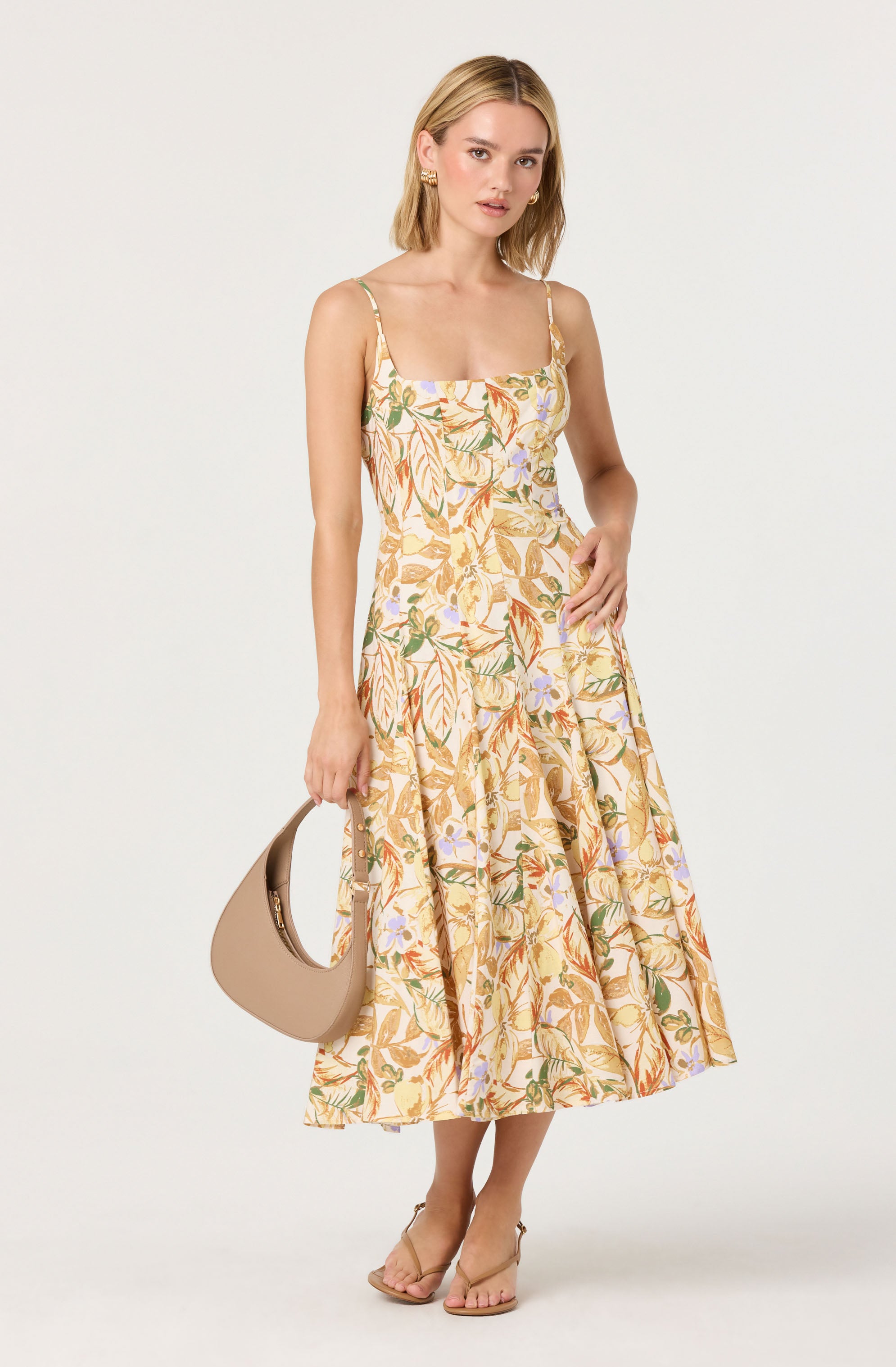 A woman is wearing a sleeveless, midi-length dress with a floral and leaf pattern in shades of yellow, orange, green, and purple on a light background. The dress has thin spaghetti straps and a fitted bodice that flares out at the skirt. She is holding a beige, curved handbag in her right hand and wearing open-toe, ankle-strap high heels.