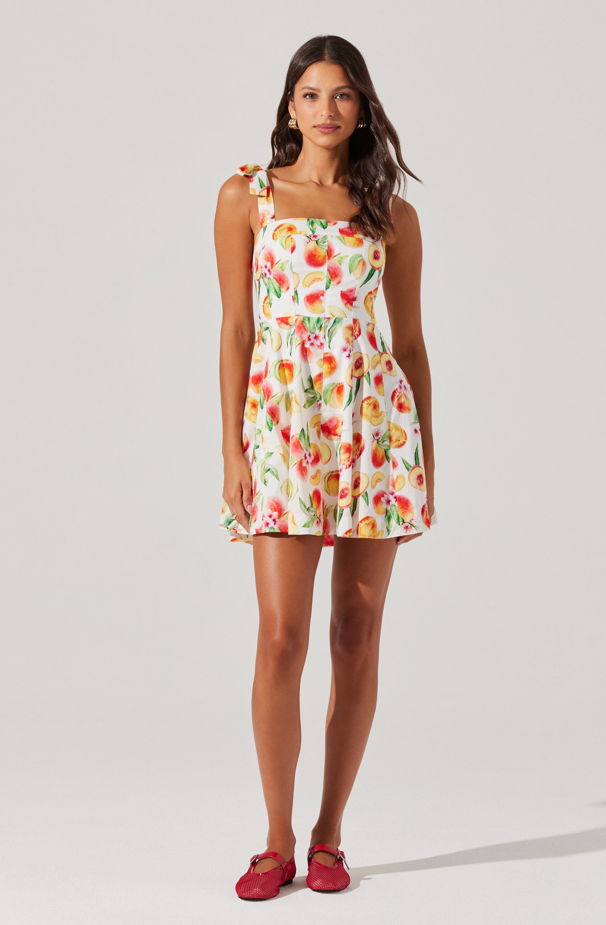 A woman is wearing a sleeveless, above-the-knee dress with a square neckline and ruffled shoulder straps. The dress features a vibrant, colorful fruit print on a white background. She has paired the dress with red shoes.