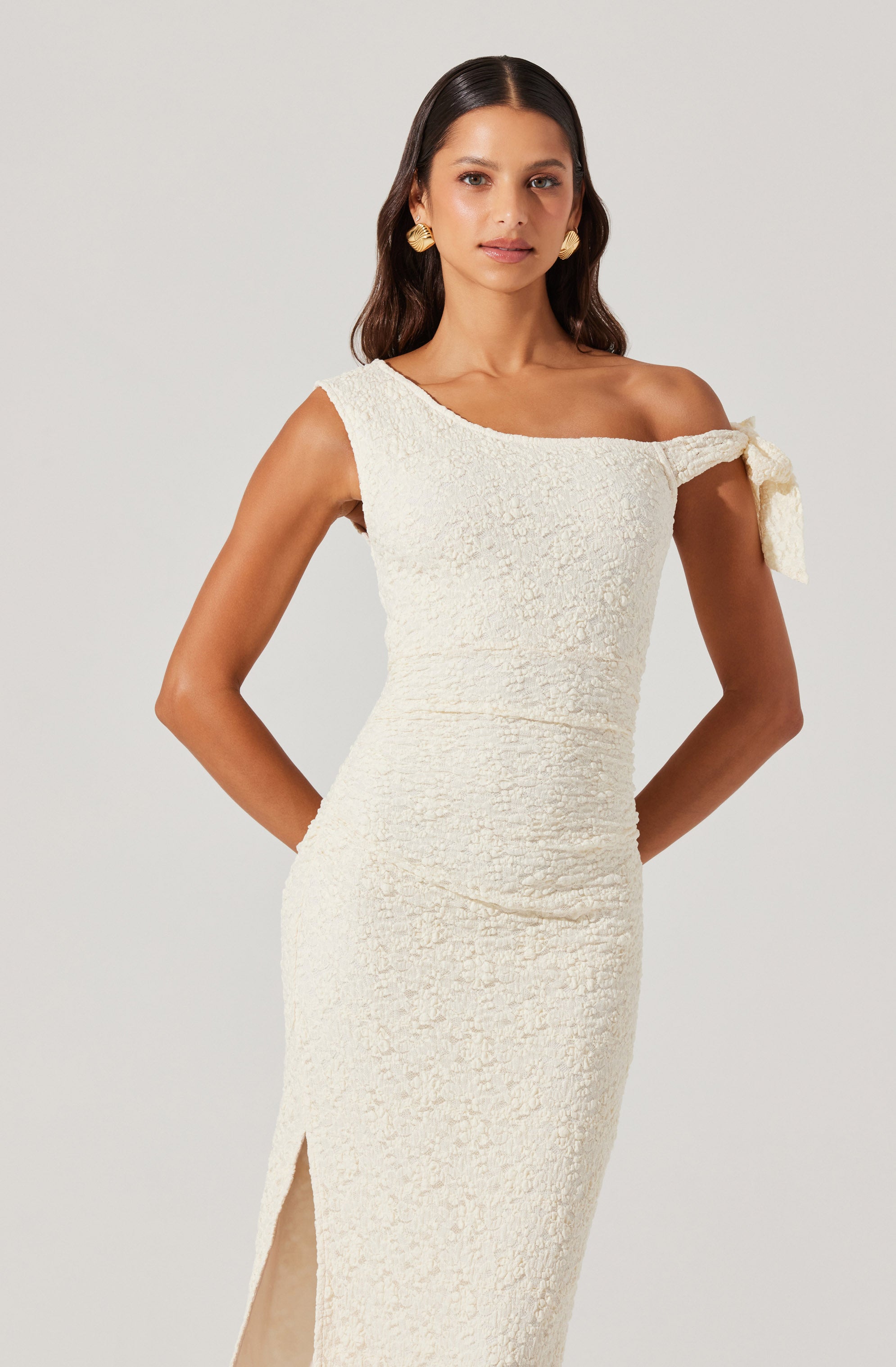 A woman is wearing a white, textured, fitted dress with an asymmetrical neckline. The dress features a single shoulder with a decorative bow on the shoulder strap and a slit on the side. She accessorizes with large, round gold earrings.