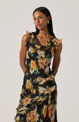 A woman is wearing a sleeveless, floral dress with ruffled shoulder details and a draped neckline. The dress features a black background with large, colorful flower prints in shades of yellow, peach, pink, and green. The fabric has a flowing, layered design with ruffles along the bodice and skirt, and a slit in the front.