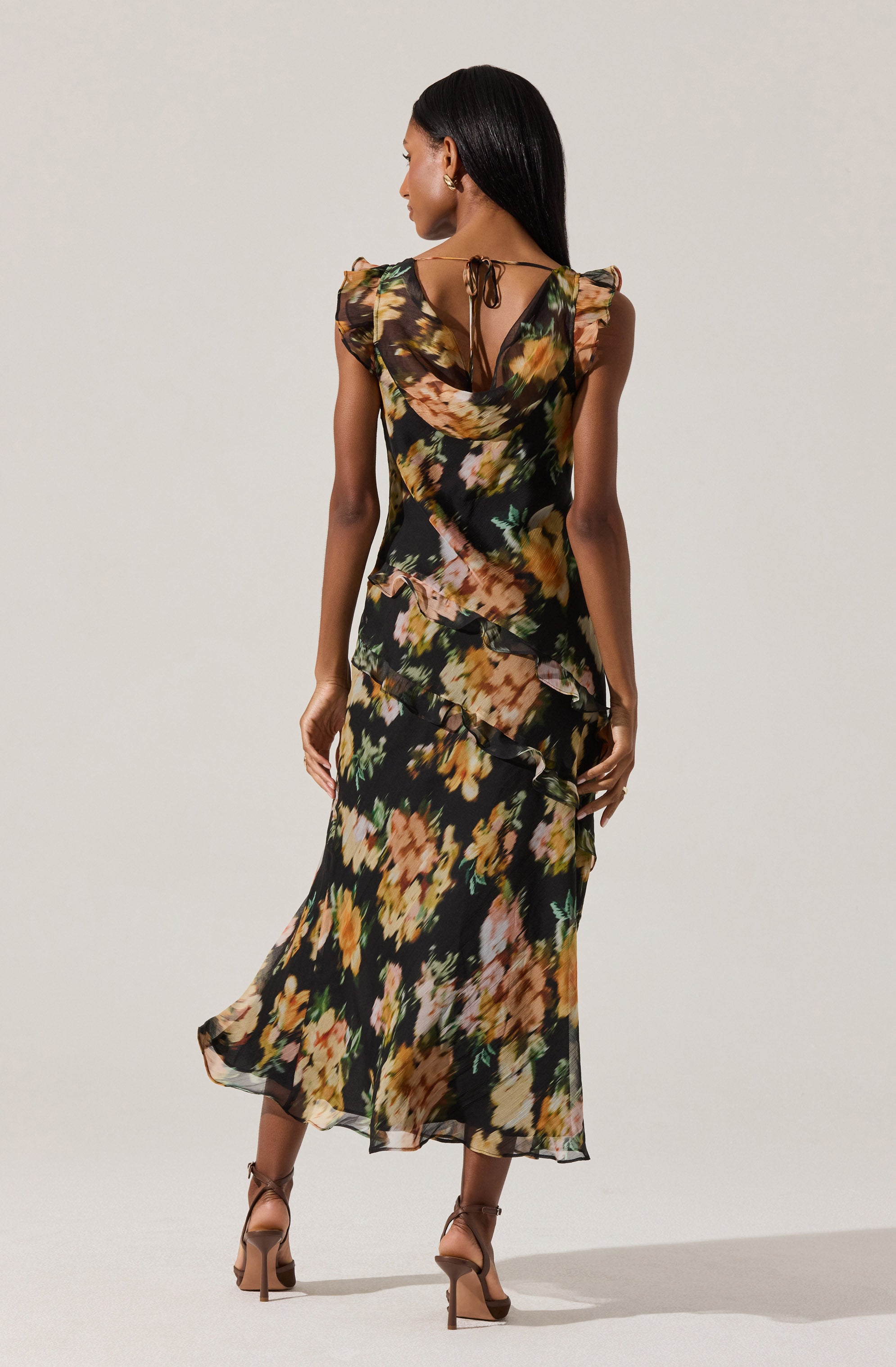 A woman is wearing a long, flowing dress with a black background and an abstract floral print in shades of yellow, orange, pink, and green. The dress features ruffled details along the sides and has shoulder pads. It has a tie at the back of the neck, creating a keyhole opening at the upper back. The fabric appears lightweight and semi-sheer. She is also wearing high-heeled sandals in a nude tone.