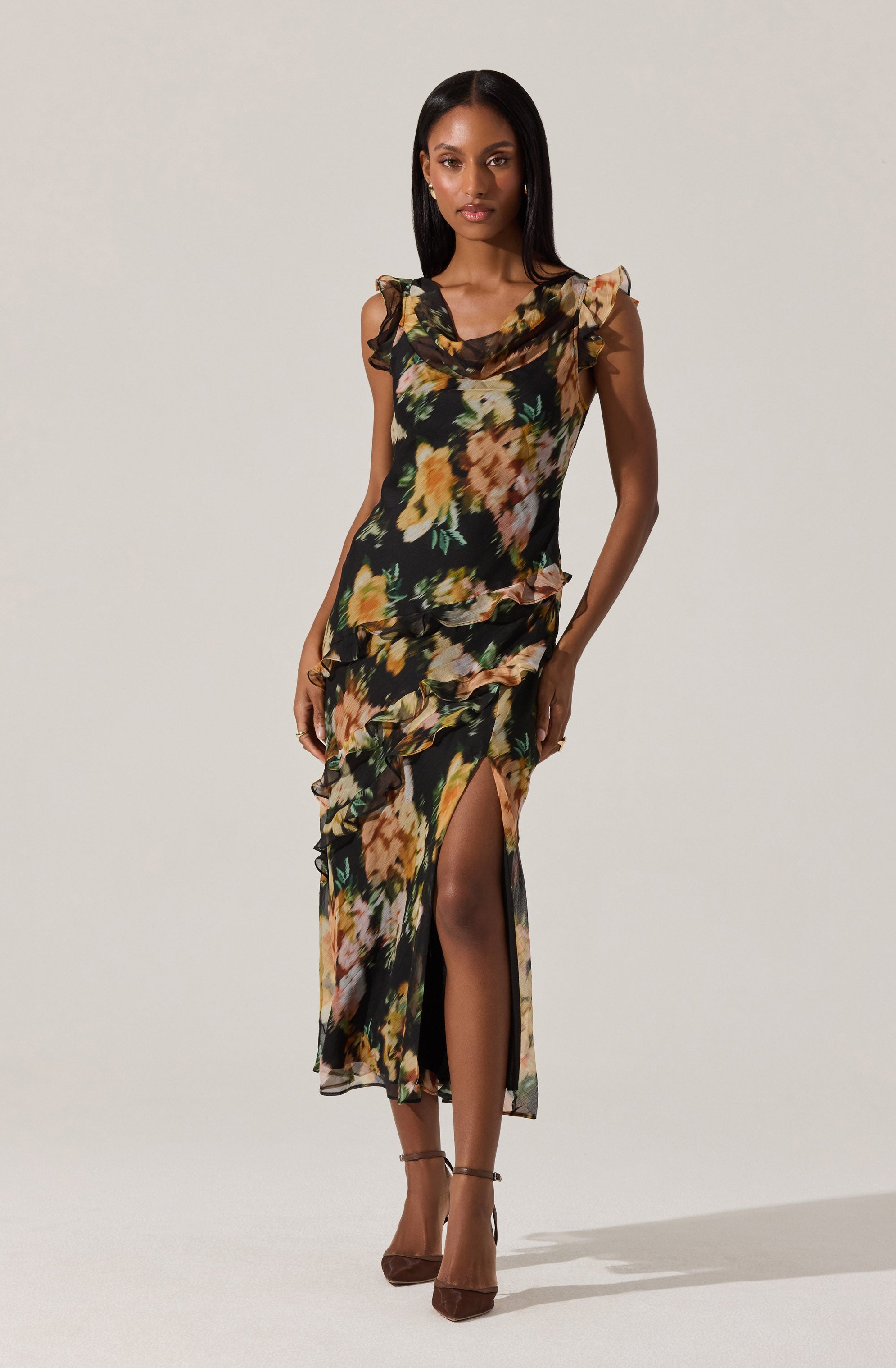 The dress is a sleeveless, floor-length gown with a black background and a floral pattern featuring shades of yellow, orange, green, and beige. It has ruffled accents along the shoulders and down the front, and a high slit on one side revealing the leg.