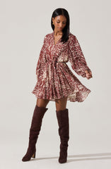 A woman is wearing a long-sleeved, V-neck dress with a leopard print pattern in shades of brown and beige. The dress has a gathered waist with a matching fabric belt, creating a slightly flared, flowy silhouette. She pairs the dress with knee-high, dark brown suede boots with a chunky heel.