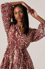 The dress features a loose, flowing fit with a collared neckline and long, slightly puffed sleeves that gather at the cuffs. It is printed with a bold, abstract animal print pattern in shades of reddish-brown and beige. The dress has a waist tie that cinches at the waist, creating a feminine silhouette.