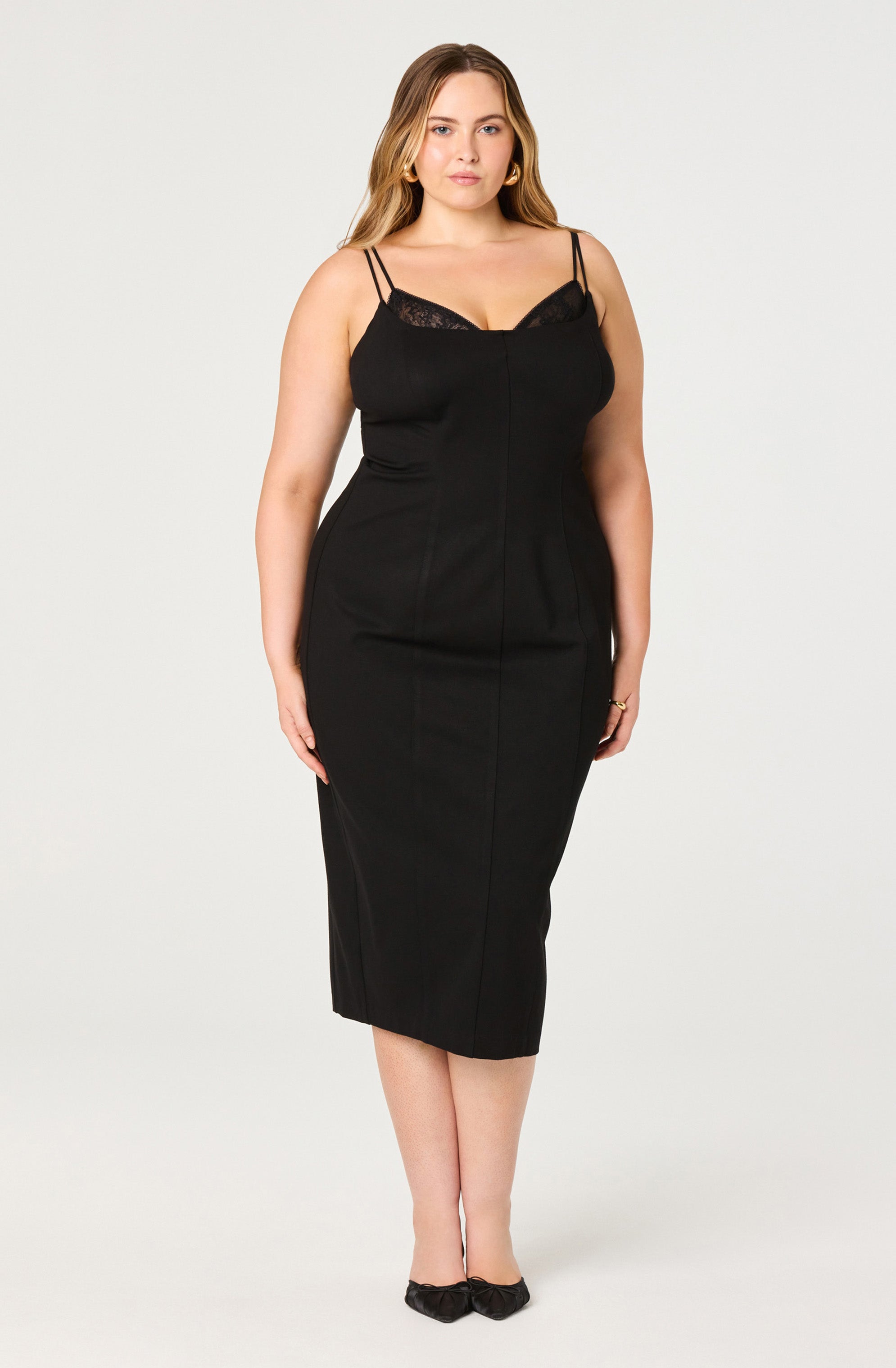 A woman is wearing a black sleeveless dress with double spaghetti straps and lace detail at the bust. The dress is fitted, midi-length, and has a sleek, elegant style. She is also wearing black shoes with a small bow detail.
