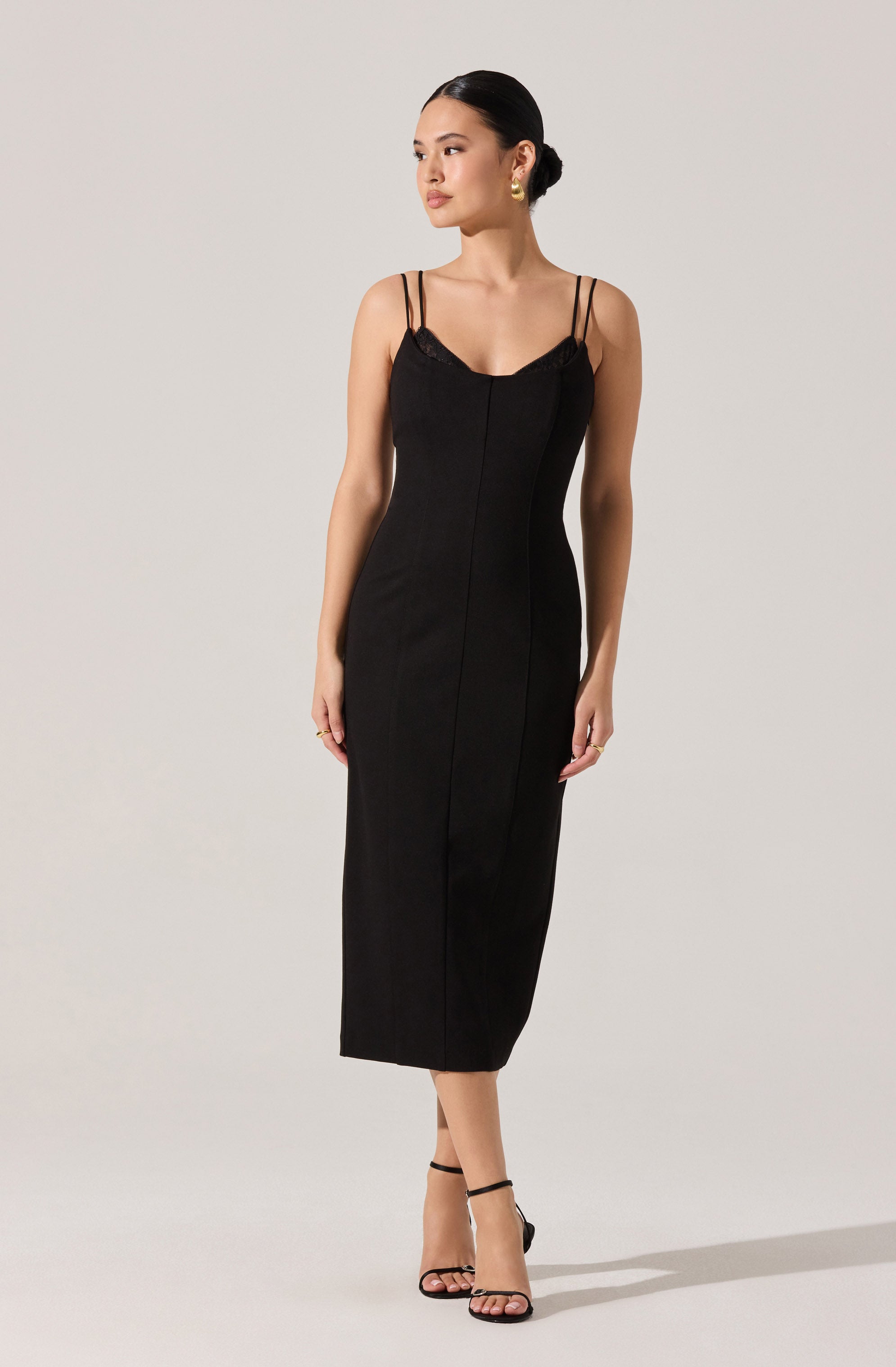 A woman is wearing a sleeveless black dress with thin, double spaghetti straps. The dress features a fitted bodice with a slight sweetheart neckline and a lace trim along the top edge. The dress has a sleek, midi-length design that hugs her figure down to the hem. She is also wearing black high-heeled sandals with ankle straps.