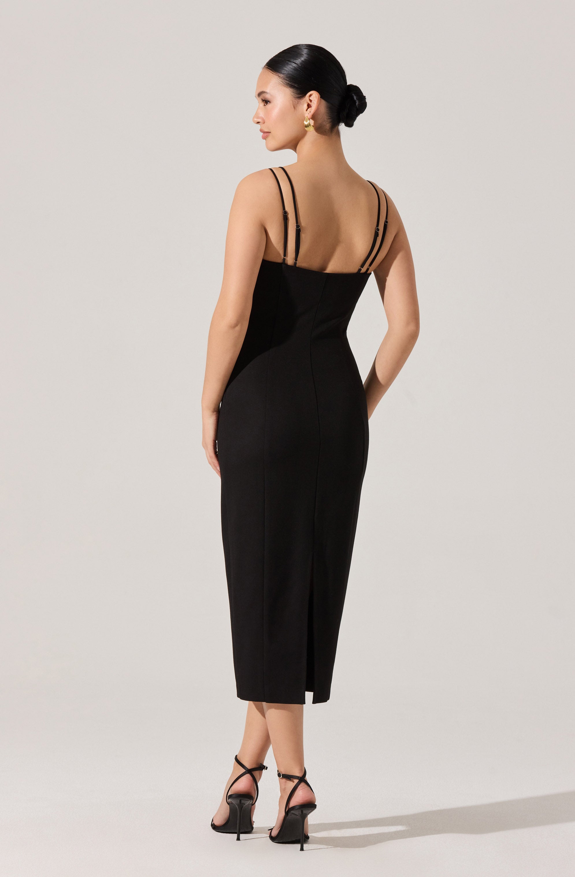 A woman is wearing a fitted, sleeveless black dress that extends below the knees and features a slit at the back. The dress has thin double straps across the shoulders, and she is accessorizing with gold earrings and black high-heeled sandals with ankle straps. Her hair is styled in a sleek bun.