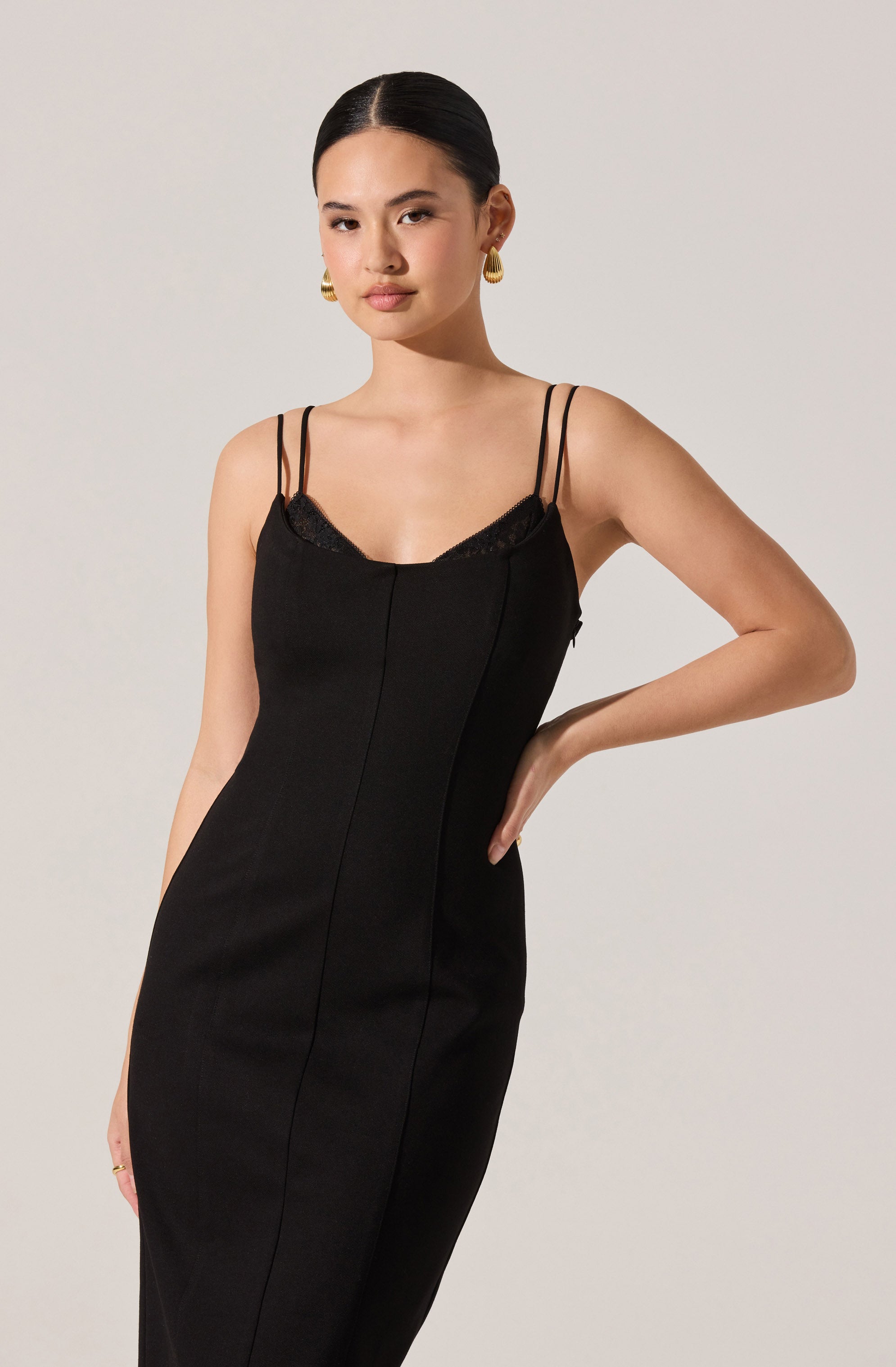 A woman wearing a black, fitted dress with thin double shoulder straps. The dress has a slight V-neckline with lace trim detail. She is accessorized with large, gold hoop earrings.