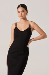 A woman wearing a black, fitted dress with thin double shoulder straps. The dress has a slight V-neckline with lace trim detail. She is accessorized with large, gold hoop earrings.