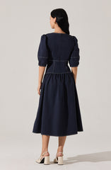 A woman is wearing a navy blue dress with puffed three-quarter-length sleeves, a fitted waist with white topstitching details, and a gathered, full-length skirt. She pairs the dress with light-colored, high-heeled sandals that have ankle straps.