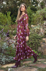 A woman is wearing a long, flowy dress with a deep V-neckline and long, semi-transparent puff sleeves. The dress is burgundy with a floral pattern featuring lavender and green leaves. She pairs the dress with dark purple high-heeled boots.