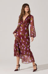 A woman is wearing a long, flowy dress with a deep V-neckline. The dress is sheer and has a maroon base color with a floral pattern of purple flowers and green leaves. The dress features long, slightly puffed sleeves gathered at the wrists and has a high slit on one side.