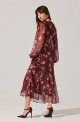 A woman is wearing a long, sheer burgundy dress with a floral print of purple flowers and green leaves. The dress has long, puffy sheer sleeves, a fitted waist, and a flowy skirt that falls to her ankles. She is also wearing burgundy high-heeled shoes with ankle straps.