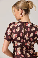 Back view of a woman wearing a dark-colored dress with a floral pattern featuring pink, white, and beige flowers. The dress has short sleeves and a vertical zipper running down the back. The fabric appears lightweight and slightly sheer. The woman has blonde hair styled in a low, loose bun and is wearing large, dangling earrings.