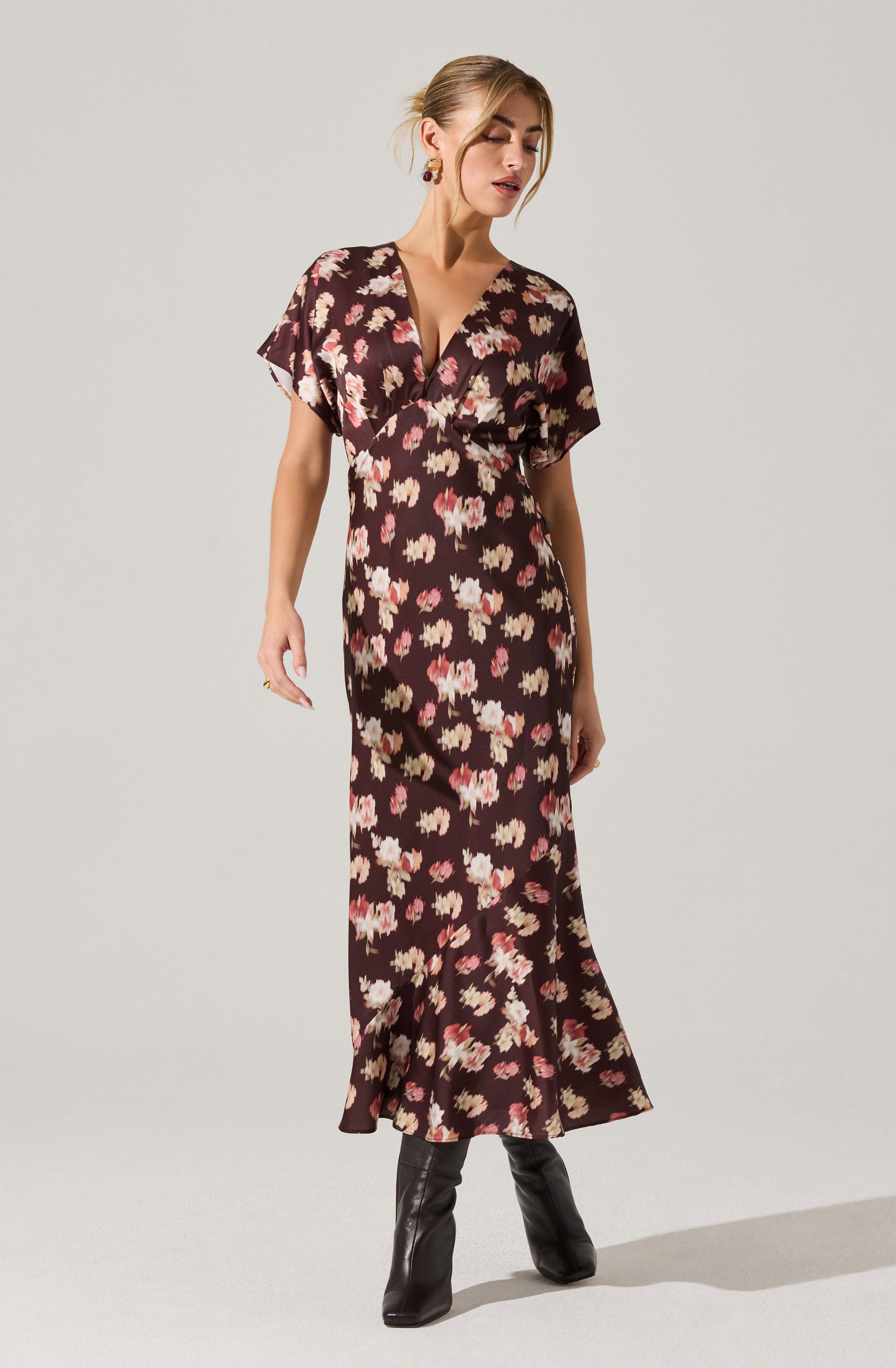 A woman is wearing a long, dark brown dress with a floral pattern featuring pink, cream, and light brown flowers. The dress has short, wide sleeves and a V-neckline. She has paired the dress with black, knee-high boots.