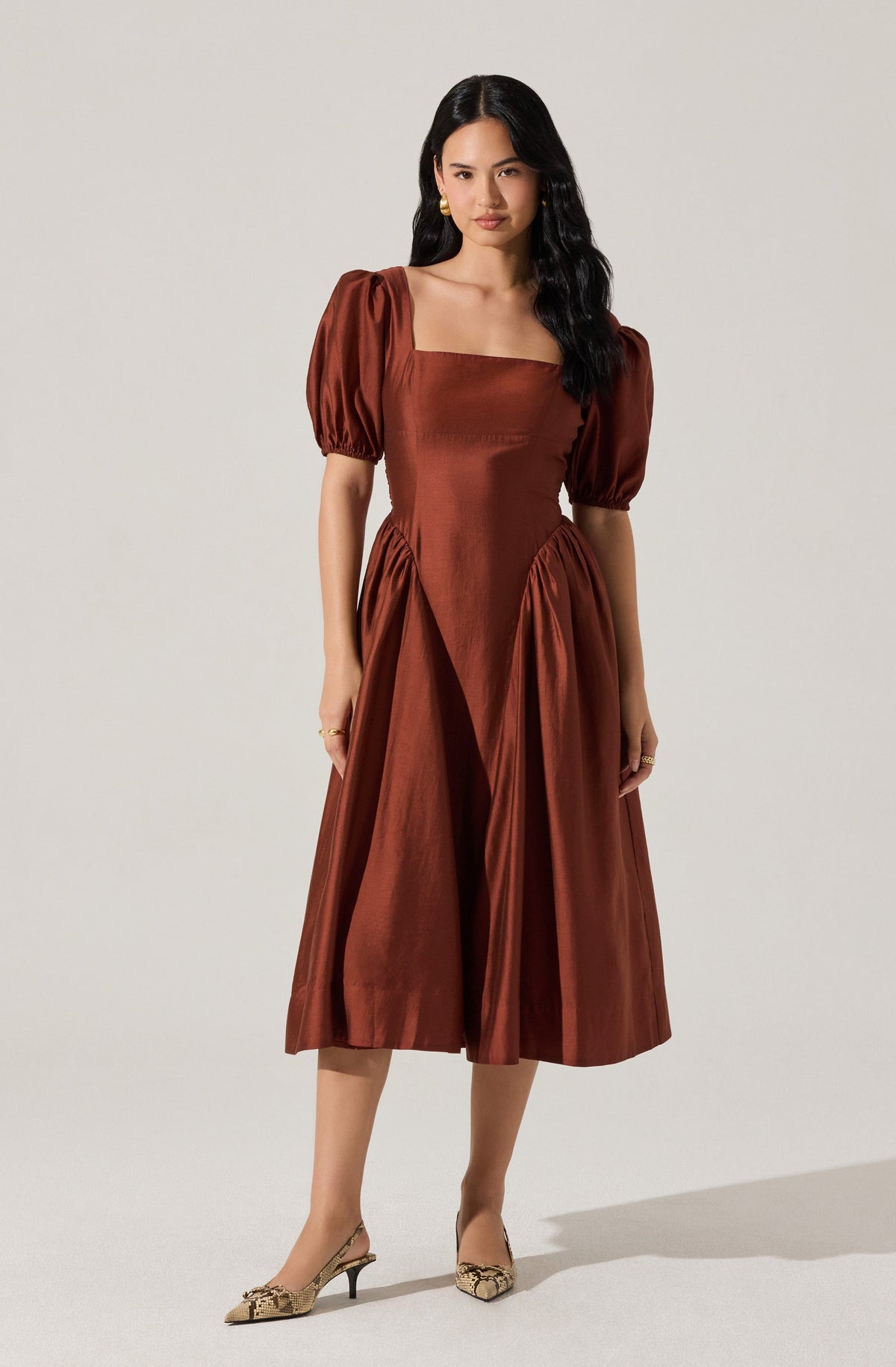 A woman wearing a mid-length, rust-colored dress with puffed short sleeves, a square neckline, and pleated details on the skirt. She is also wearing beige heels with a animal print pattern.