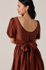 A woman is wearing a bronze-colored dress made of a shiny, satin-like fabric. The dress features puffed short sleeves, a wide scoop neckline at the back, and a large, tied bow at the waist. The skirt is gathered and falls in soft pleats from the waist. She has dark, shoulder-length hair and is accessorized with large, gold hoop earrings.