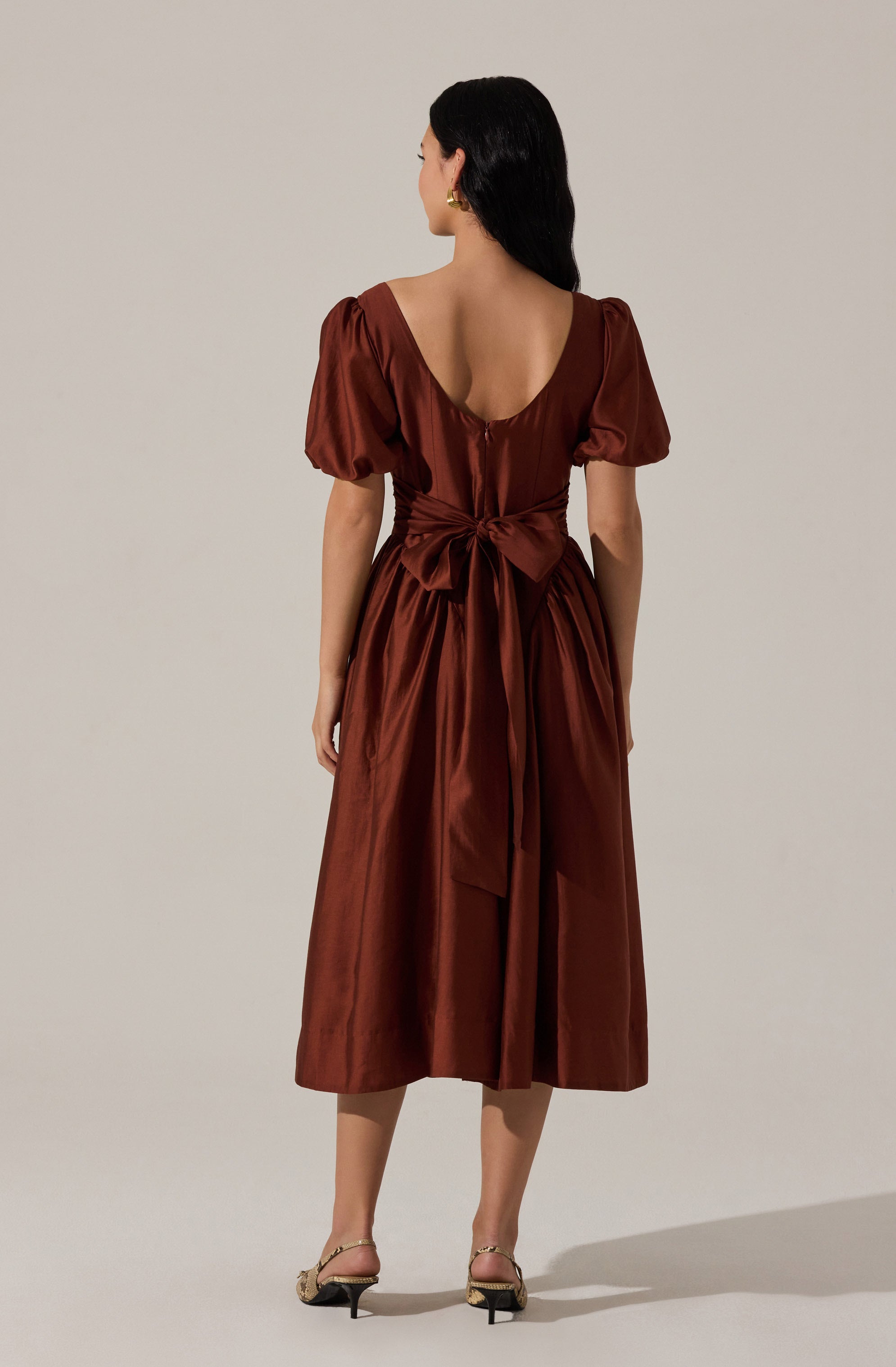 A woman is wearing a midi-length, satin dress in a rich, rust-brown color. The dress features short, puffed sleeves, a deep scoop back neckline, and a wide sash tied into a large bow at the waist. The fabric has a shiny, smooth appearance.