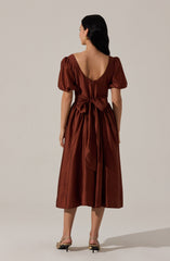 A woman is wearing a midi-length, satin dress in a rich, rust-brown color. The dress features short, puffed sleeves, a deep scoop back neckline, and a wide sash tied into a large bow at the waist. The fabric has a shiny, smooth appearance.