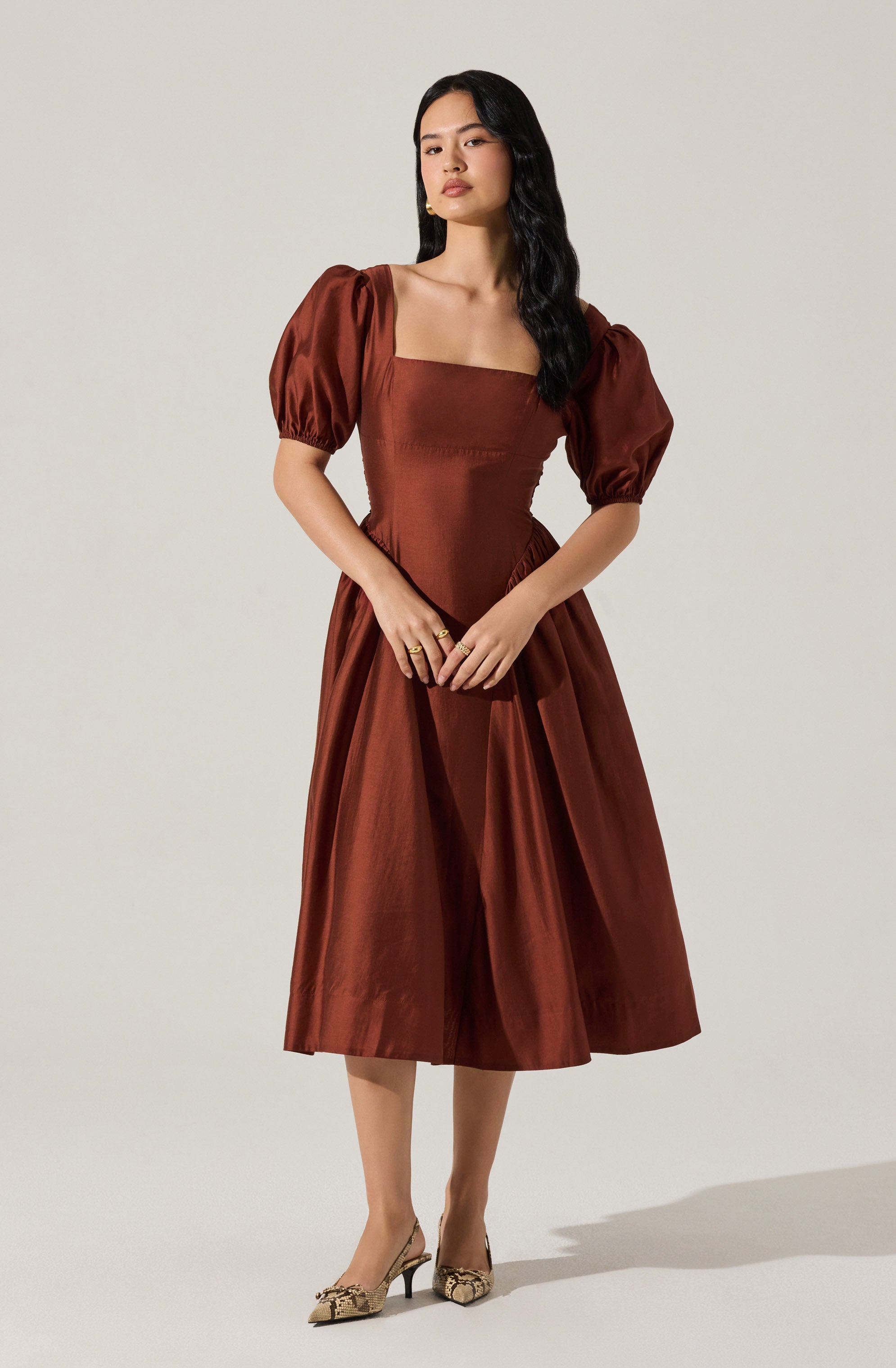 A woman is wearing a rust-colored dress with a fitted bodice and square neckline. The dress has puffed short sleeves that gather at the elasticized cuffs, and a full, midi-length skirt with pleats. She is also wearing beige high-heeled shoes with a snakeskin pattern.