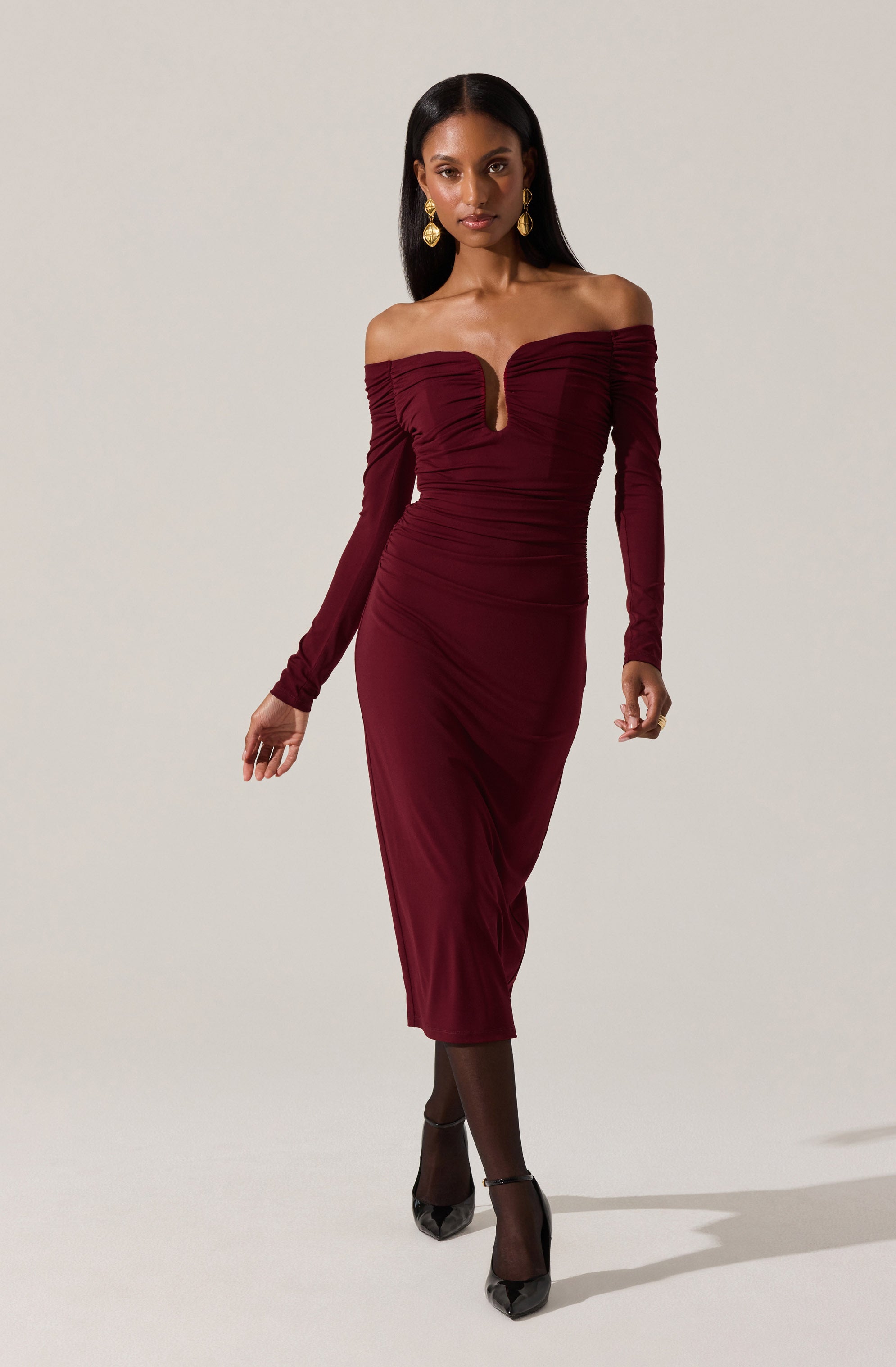 A woman wearing an elegant, off-the-shoulder, long-sleeve burgundy dress with ruched detailing across the bodice and sleeves. The dress has a fitted silhouette that falls to mid-calf length. She pairs the outfit with black tights, black heels, and gold earrings.