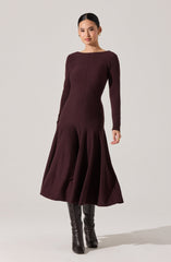 A woman is wearing a long, dark burgundy dress with a fitted bodice and long sleeves. The dress has a subtle vertical center seam and a flared, pleated skirt that falls to mid-calf length. She is also wearing black knee-high boots.