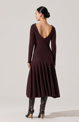 A woman is wearing a long, burgundy dress with a fitted bodice that has a deep V-back and long sleeves. The skirt of the dress features pleats that add volume and movement, ending at mid-calf length. She is also wearing tall, dark-colored high-heeled boots.