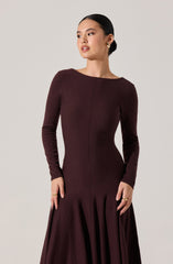 A woman is wearing a fitted, long-sleeved, dark burgundy dress with a round neckline. The dress features a subtle vertical seam down the front and a pleated, flowing skirt that extends to the floor. She has her hair tied back and is accessorized with gold hoop earrings and gold rings.