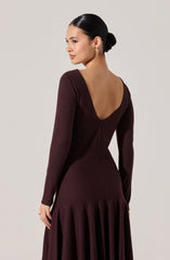 A woman is wearing a long, fitted, deep burgundy dress with long sleeves. The dress features a low, rounded back neckline, exposing her upper back. The skirt of the dress is flowing with gathered fabric at the waist. The fabric appears to be smooth and slightly textured.