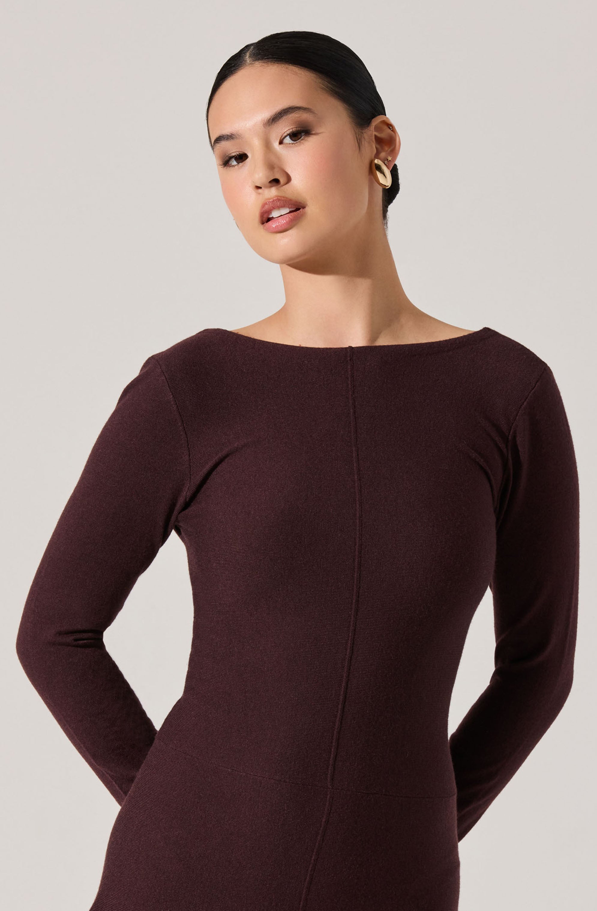 A woman is wearing a long-sleeved, form-fitting dress in a dark burgundy color. The dress has a simple, elegant design with a round neckline and visible seam details running vertically down the front. She has dark hair pulled back and is accessorizing with large, rounded gold earrings.