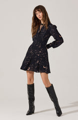 A woman is wearing a dark-colored dress with a celestial pattern featuring moons, stars, and clouds in light pastel shades. The dress has long sleeves, a fitted waist, and a slightly flared skirt that falls above the knees. She pairs the dress with tall, black, shiny knee-high boots.