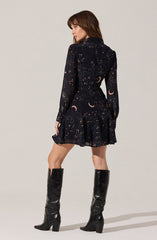 A woman is wearing a dark dress with a celestial pattern featuring moons and stars. The dress has long sheer sleeves, a fitted waist, and a flared skirt. She pairs the dress with tall, shiny black knee-high boots with block heels.