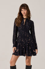 A woman is wearing a dark navy dress adorned with a celestial print featuring moons, stars, clouds, and sparkles. The dress has long sleeves, a fitted waist, and a short, flared skirt. It also features a high neckline with a keyhole opening tied with a black ribbon bow.