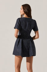 A woman is wearing a dark, textured, short-sleeved dress with a subtle shimmer or sparkle. The dress has a fitted bodice that cinches at the waist and a flared skirt that falls above the knees. The fabric appears to have a pattern of tiny, light-colored specks evenly distributed throughout.