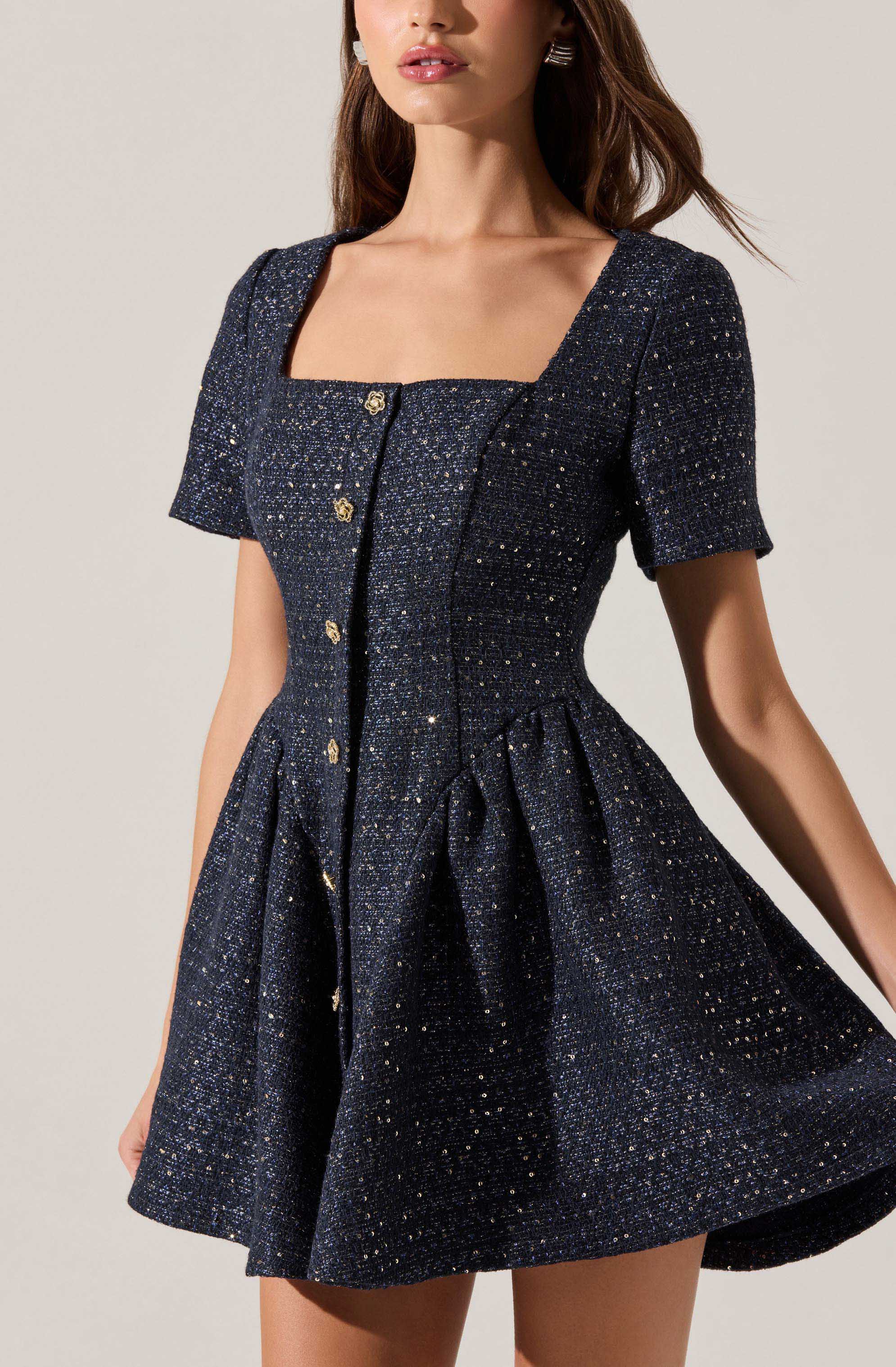 A woman is wearing a dark-colored, sparkly, textured dress with short sleeves and a square neckline. The dress features a fitted bodice with decorative buttons down the front and a flared skirt with pleats. The fabric has a shimmering appearance with small, glitter-like details throughout.